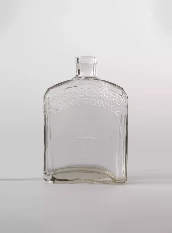 Przezroczysta szklana butelka. Dolna część jest wysoka, prostokątna, lekko zaokrąglona u góry. Szyjka butelki jest krótka.