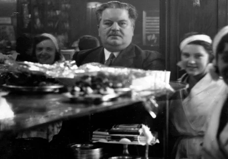 Czarno-biała fotografia wnętrza kawiarni. Na ladzie na szklanych półkach ciasta. Za ladą tęgi mężczyzna z wąsami i dwie kelnerki w białych fartuszkach.