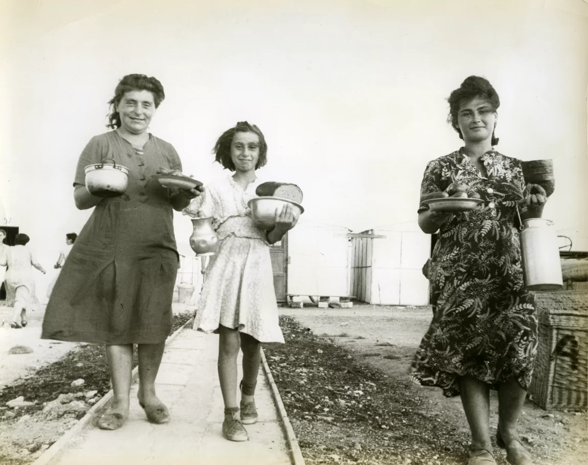 Czarno-biała fotografia. Chodnikiem idą dwie kobiety i dziewczynka. Niosą naczynia z jedzeniem.