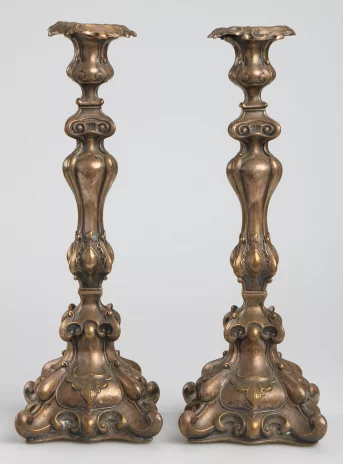 Dwa takie same świeczniki. Są pojedyncze, wysokie i mosiężne. Ich nóżka jest dekoracyjnie uformowana.