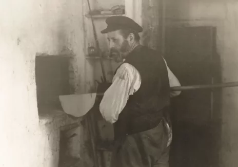 Kitchen interior. A middle-aged man at the bread oven. The man is dressed in a white shirt and a dark vest. On his head he has a cap with a small visor.