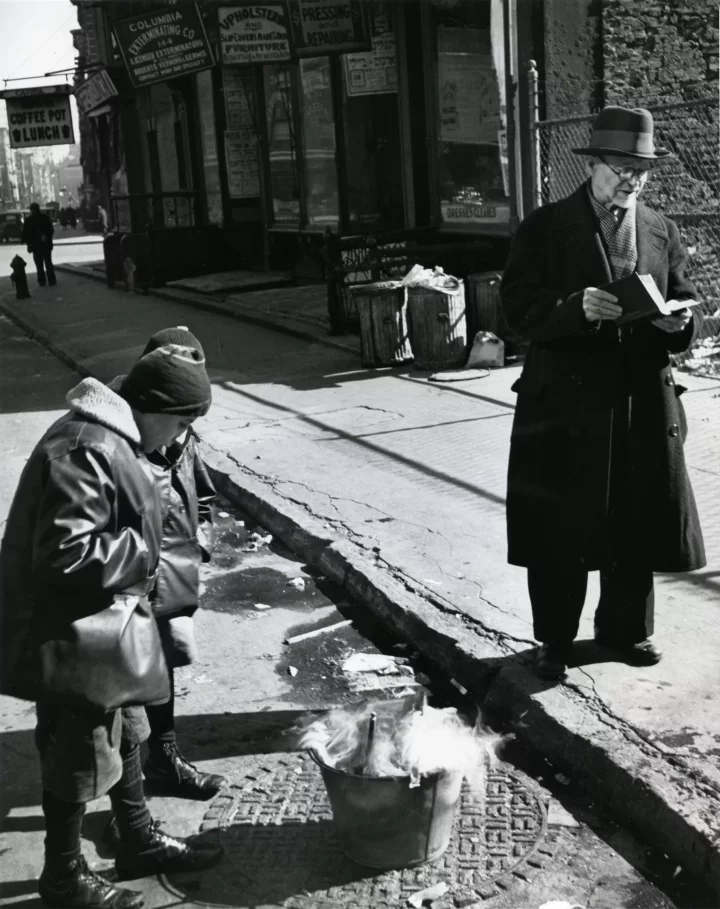 Black and white photo. On the sidewalk stands a man in a dark coat and hat. In his hands he holds an open prayer book. A woman smokes in a bucket chamec.